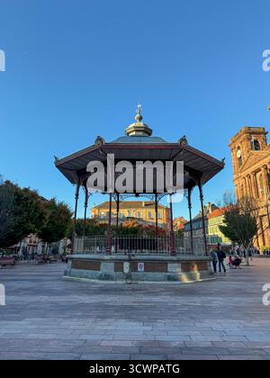 Verzierte gusseiserne Bühne auf dem zentralen Platz von Belfort mit historischer Kirche und Uhrenturm im Hintergrund unter der warmen Nachmittagssonne Stockfoto