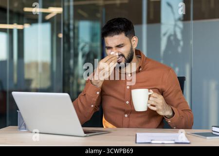 Junge Profis am Schreibtisch, die mit Kälte oder Allergien kämpfen, während sie am Computer arbeiten, warme Tasse und Taschentücher halten und Müdigkeit und Unwohlsein während des Arbeitstages zeigen Stockfoto