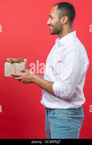 Lächelnder Mann in weißem Hemd, der Geschenkbox mit Geschenkband-Schleife auf rotem Hintergrund hält Stockfoto