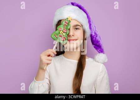 Mädchen mit weihnachtlichem Hut, der baumförmige Süßigkeiten hält. Festliches Mädchen in lila Weihnachtsmütze, das Weihnachtsbaumbonbons vor ihrem Auge auf Pastell hält Stockfoto