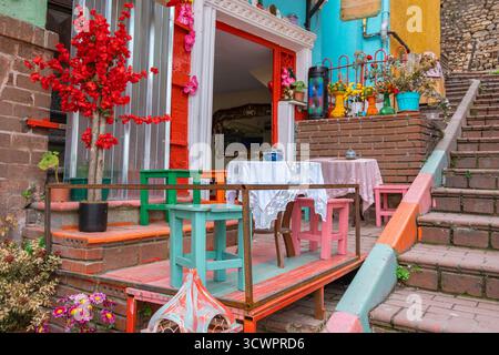 Farbenfroher Café-Tisch und Stühle im Freien mit Vintage-Holzhockern und Blumen auf gemütlicher Backsteinterrasse. Charmantes rustikales Straßencafé mit kleinem Holz Stockfoto