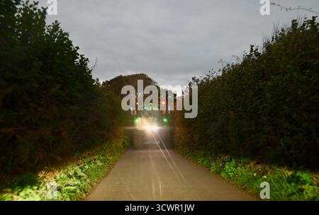 Ein Traktor mit hellen Scheinwerfern blockiert die Straße auf einer schmalen Landstraße in devon england Stockfoto