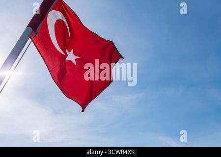 Rote türkische Flagge mit weißem Stern und Sichel fliegt auf Fahnenmast, sonniger Tageshintergrund. Winkend im Wind vor hellblauem Himmel, nationales Symbol Stockfoto