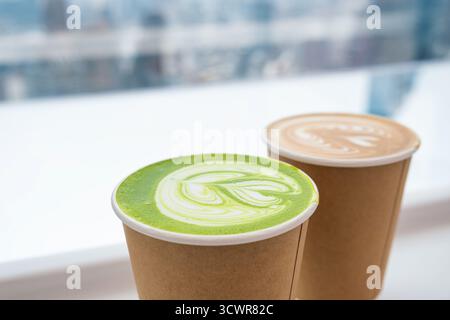 Zwei Tassen zum Mitnehmen mit Matcha Latte und Kaffee Latte mit künstlerischen Schaummustern in der Nähe des hellen Fensters im Café. Nahaufnahme. Flach Stockfoto