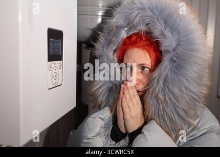 Im Inneren wärmt eine Frau mit einem großen Kapuzenmantel ihre Hände. Sie steht in der Nähe einer weißen Heizungsanlage und sieht tagsüber kalt in ihrem Haus aus. Stockfoto