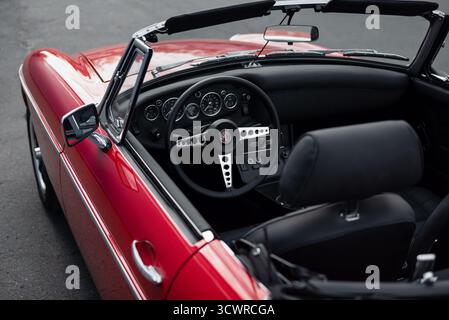 Innenraum des roten MGB Classic Roadsters. 1960er Jahre MG Sportwagen mit schwarzem Lederinnenraum, Asphaltstraße im Hintergrund. Stockfoto