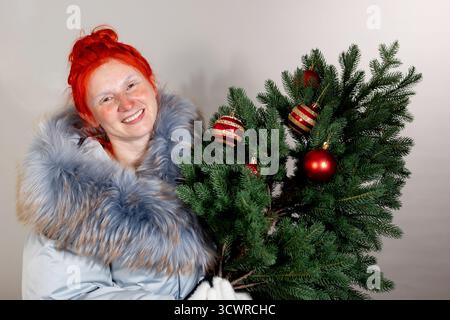 Frau im Wintermantel lächelt, nachdem sie draußen in der Kälte war. Sie hält einen kleinen grünen Weihnachtsbaum, der mit roten und goldenen Ornamenten verziert ist. Stockfoto