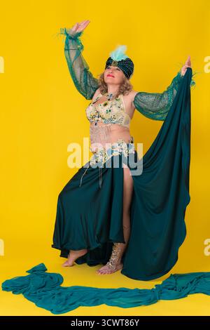 Glückliches reifes Damen-Modell mit handerhöhtem Anzug im grünen östlichen Tanzbühnenkostüm - Turban, BH-Oberteil und langer Rock mit Schlitz, Bewegungen und Stockfoto