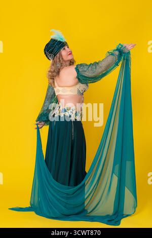 Kurviges, reifes Model für Erwachsene Frauen, das in einem grünen Kostüm der östlichen Tanzbühne trägt – Turban, BH-Oberteil und langer Rock mit Pareo, posiert und zieht ein Stockfoto