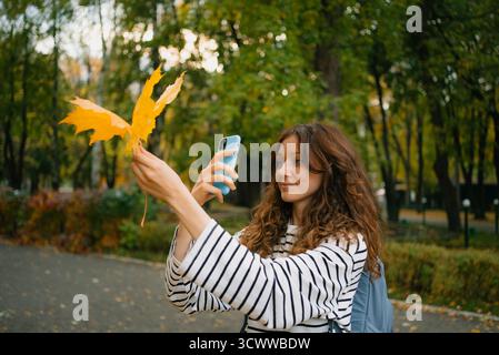 Frau mit einem hellgelben Ahornblatt, die mit einem Smartphone seine leuchtende Farbe und Textur in einem üppigen Herbstpark fotografiert und die Herbstsaison festgehalten Stockfoto