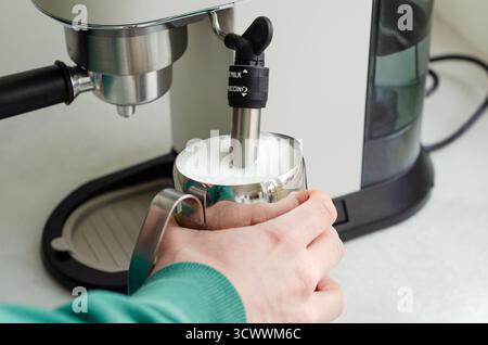Gemütlicher Morgen mit männlichen Händen, die Milch im Milchkrug aufschäumen, mit einer Kaffeemaschine für Cappuccino zu Hause Stockfoto