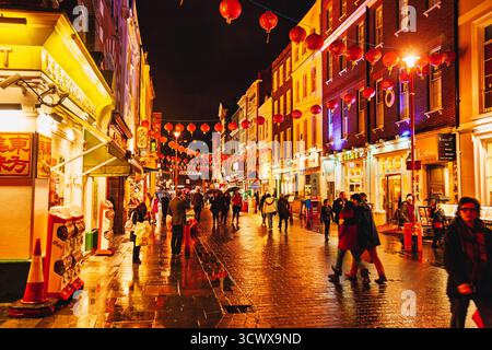 London, England – 30. Oktober 2012: Nachtblick auf Chinatown mit beleuchteten roten Laternen, die über der nassen Straße hängen, spazierenden Menschen und neonbeleuchtetem Restaurant Stockfoto