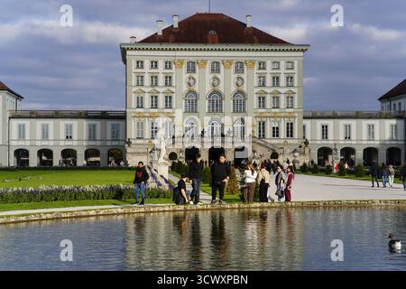 Besucher genießen das nachmittags Sonnenlicht am reflektierenden Teich vor dem Schloss Nymphenburg in München, Deutschland. Schloss Nymphenburg, München, Deutschland Stockfoto
