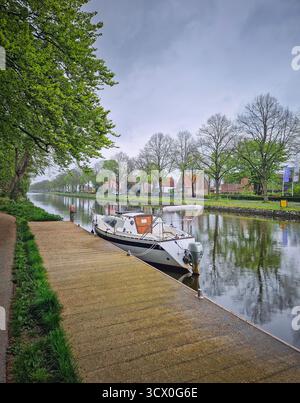 Eine ruhige Kanalszene an einem bewölkten Tag mit einem kleinen Boot, das an einem Dock im Vordergrund verankert ist. Üppiges Grün im Frühjahr und friedliche Atmosphäre Stockfoto
