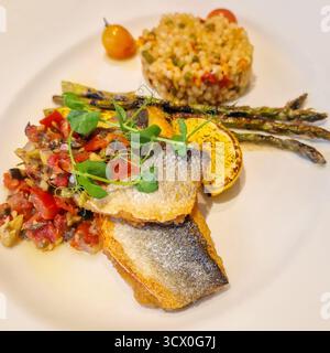 Ein lebhaftes Gericht mit knusprigen Seebarschfischfilets, serviert mit einer lebhaften Tomaten- und Oliven-Salsa-Relief, gegrillten Spargeln und einer Schaufel davon Stockfoto