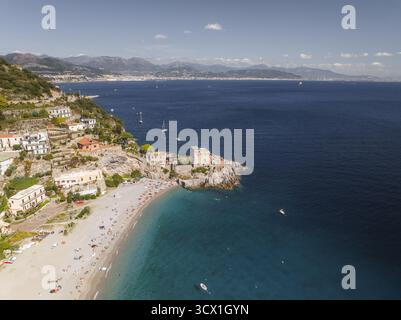Aus der Vogelperspektive auf einen sonnengeküssten Strand, der auf das azurblaue Meer trifft, eingerahmt von Terrassengebäuden, die sich an grünen Hängen schmiegen, Erchie, Amalfiküste, Italien. Stockfoto