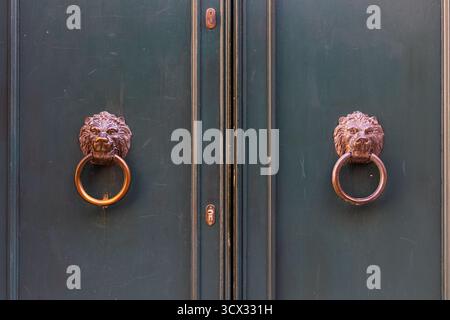 Antike Löwenkopf-Türklopfer an der Doppeltür. Nahaufnahme von zwei verzierten Bronze Löwenkopf-Klopfern, die an einer dunklen Holzdoppeltür angebracht sind, symbolisieren Stockfoto