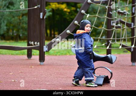 Der verspielte Junge in blauen wasserdichten Overalls steht neben einem Frühlingsspielzeug auf dem Spielplatz und genießt an einem kühlen Herbsttag Spaß im Freien und Fantasie. Stockfoto