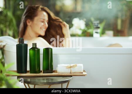 Eine lächelnde junge Frau entspannt sich in einer weißen Badewanne; natürliche Badeprodukte auf einem Holztablett stehen im Fokus. Das helle, ruhige Badezimmer mit verschwommenem Grün Stockfoto