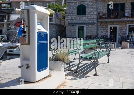 Ein moderner öffentlicher Ladestation mit mehreren Steckdosen und Kabeln befindet sich neben einer blauen Tafel vor einem Steingebäude und städtischen Sitzgelegenheiten. Ideal für Stockfoto