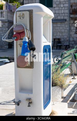 Ein moderner öffentlicher Ladestation mit mehreren Steckdosen und Kabeln befindet sich neben einer blauen Tafel vor einem Steingebäude und städtischen Sitzgelegenheiten. Ideal für Stockfoto