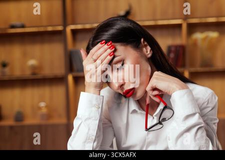 Nahaufnahme einer müden Frau, die ihren Kopf mit einer Brille in der Hand hält, was Migräne und Stress in Verbindung mit Triggern und Arbeitsdruck veranschaulicht Stockfoto