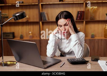 Müde Büroangestellte reiben die Bügel und halten die Brille neben dem Laptop, um Migräne, Bildschirmmüdigkeit und Stress bei der Nachtarbeit an einem modernen Arbeitsplatz zu veranschaulichen Stockfoto