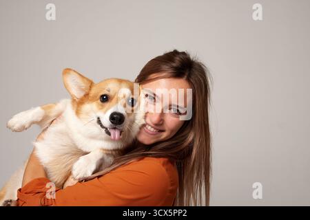 Lächelnde junge Frau, die ihren Corgi in einem Studio kuschelt. Porträt liebevoller Haustierbesitzer mit Kopierraum für Haustierpflege oder Lifestyle-Themen. Stockfoto