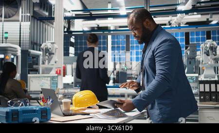 Ein afroamerikanischer Branchenexperte überprüft die Photovoltaik-Montagelinie und überprüft die Produktion, um die Effizienz im Solarenergiegeschäft zu gewährleisten. Junger Ingenieur in einer Fabrik. Kamera A.. Stockfoto