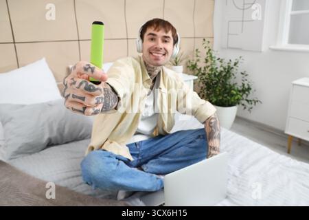 Junger gutaussehender Mann in Kopfhörern mit elektronischer Wegwerfzigarette, der Laptop auf dem Bett zu Hause benutzt Stockfoto