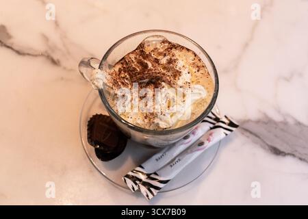 Genf, Schweiz - 15. Oktober 2024: Kaffee mit Milch und Schlagsahne in einer transparenten Tasse auf einem weißen Tisch im Restaurant. Stockfoto