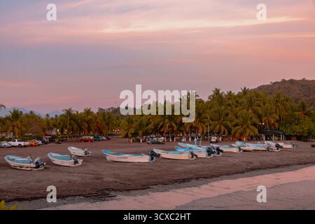 Am Playa Linda in Ixtapa, Mexiko, liegt eine Reihe farbenfroher Fischerboote auf dem Sand, während die Sonne über dem Pazifik untergeht. Stockfoto