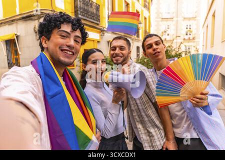 Eine Gruppe verschiedener junger Leute lächelt und feiert die Bewegung des Schwulen-Stolzes, während sie eine Regenbogenfahne, ein Megaphon und einen bunten Handfan in einer Stadt halten Stockfoto