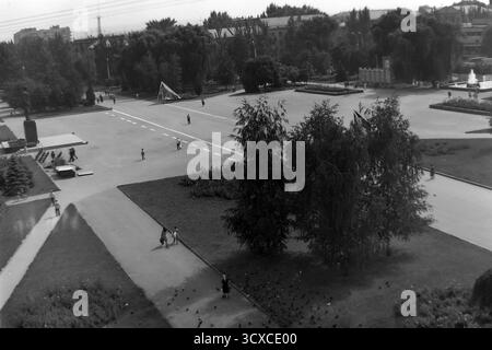 Ein Hochwinkel-Archivfoto aus den 1980er Jahren zeigt den weitläufigen Platz der Oktoberrevolution (heute Soborna-Platz) in Sloviansk, Ukrainische SSR. In diesem zentralen öffentlichen Bereich befinden sich ein markantes Denkmal auf der linken Seite, Brunnen und sowjetische Flaggen. Fußgänger schlendern gemütlich über die offene Fläche, während sich im Vordergrund eine Schar Tauben versammelt, die eine lebhafte Atmosphäre schaffen. Dieses Bild zeigt eine typische Szene des täglichen Lebens und der Freizeit in einer sowjetischen Stadt, eine wertvolle historische Aufzeichnung des friedlichen Donbas vor dem Krieg Stockfoto