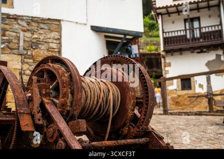 Ein rostiges altes Seil ist an einem großen Metallgegenstand in Tazones Asturien befestigt. Die Szene spielt in einem Innenhof mit einem Gebäude im Hintergrund Stockfoto