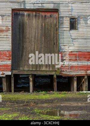 Verwittertes Wellblech-Lager mit Rost und roten Farbresten. Es ist eine große, gebrochene Holzladetür zu sehen, die von einem Metallgleis umgeben ist. Stockfoto