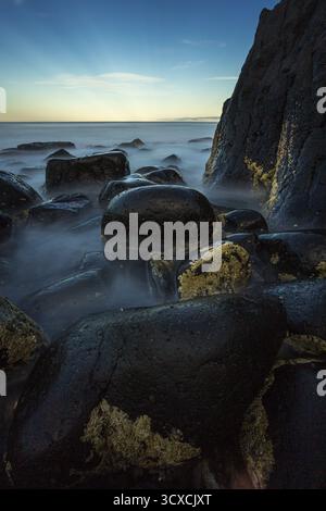 Felsige Küste in Burleigh Heads, Australien. Große, glatte, dunkle Felsbrocken werden teilweise in nebeliges Wasser getaucht, das während des Sonnenaufgangs gefangen wird. Stockfoto