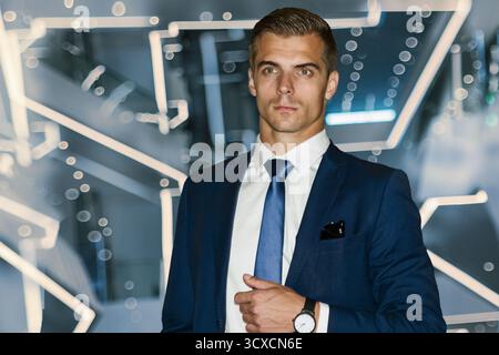 Ein männliches Model in einem dunkelblauen Anzug, weißes Hemd steht auf einem dynamischen blauen Neonhintergrund. Professioneller Manager, futuristische Neonbeleuchtung. Stockfoto