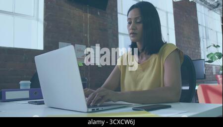 Reife asiatische Frau mit silbernem Laptop an einem modernen, offenen Schreibtisch mit Kaffeetasse Stockfoto