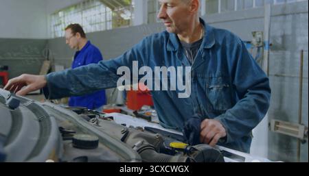 Abwischender Frontmechaniker, der in der Fahrzeugreparaturwerkstatt über den Motorraum reicht, mit geöffneter Motorhaube Stockfoto