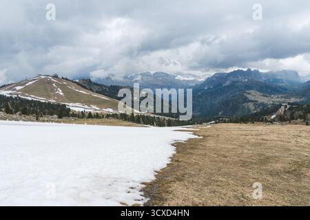 Frühlingslandschaft in den Dolomiten, Italien, mit schmelzendem Schnee auf alpinen Wiesen, sanften Hügeln und dramatischem Blick auf die Berge unter bewölktem Himmel. Stockfoto