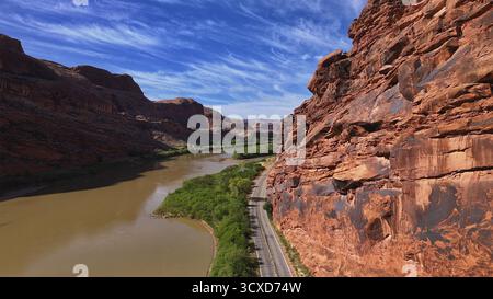 Aus der Vogelperspektive auf den schlammigen Colorado River, der sich durch die roten Felsschluchten unter einem schroffen Himmel schlängelt, entlang einer einsamen Straße, die sich am Flussufer anschmiegt, Moab, Utah, USA. Stockfoto