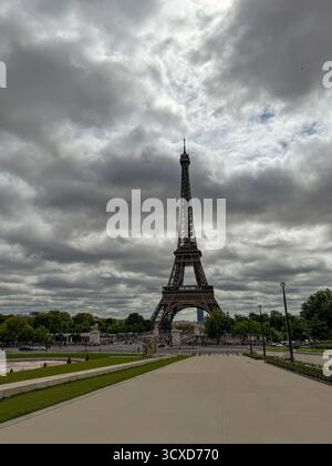 Ein flacher, vertikaler Blick auf den Eiffelturm in Paris, Frankreich, der sich über einem breiten, leeren Pfad und grünen Rasenflächen unter einem dramatisch bewölkten Himmel erhebt. Stockfoto