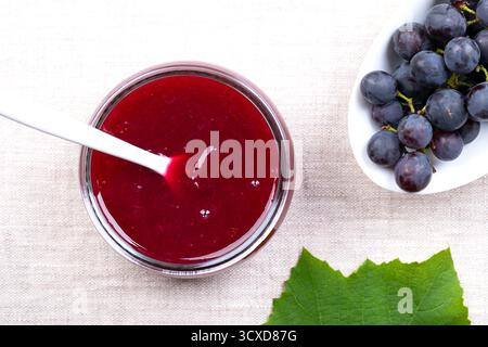 Traubenmarmelade, aus blauen Trauben, im Glas, auf Leinen. Offenes Glas mit Plastiklöffel in hausgemachte Traubenmarmelade getaucht. Stockfoto