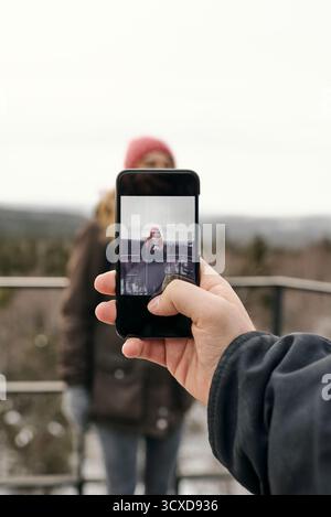 Beschneiden männliche Hand, die Handy hält und ein Foto von lächelnder Frau in Kleidung vor Winterbergen macht Stockfoto