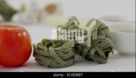 Zeigt drei Spinat-Nudelnester auf der Küchenarbeitsplatte, mit roten Tomaten, Knoblauch und Schüsseln Stockfoto