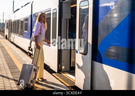 Elegante und selbstbewusste Geschäftsfrau, die am Bahnhof mit Koffer in einen Zug einsteigt. Stockfoto