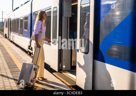 Elegante und selbstbewusste Geschäftsfrau, die am Bahnhof mit Koffer in einen Zug einsteigt. Stockfoto
