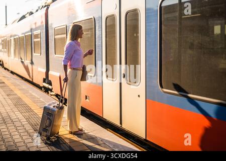 Elegante und selbstbewusste Geschäftsfrau, die am Bahnhof mit Koffer in einen Zug einsteigt. Stockfoto