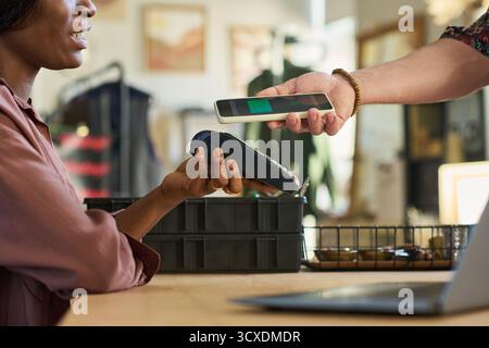 Schwarze Frau hält Zahlungsterminal, während ein kaukasischer Mann kontaktlos mit Smartphone im Second-Shop bezahlt, beide Hände sichtbar, Kleiderständer im Hintergrund Stockfoto